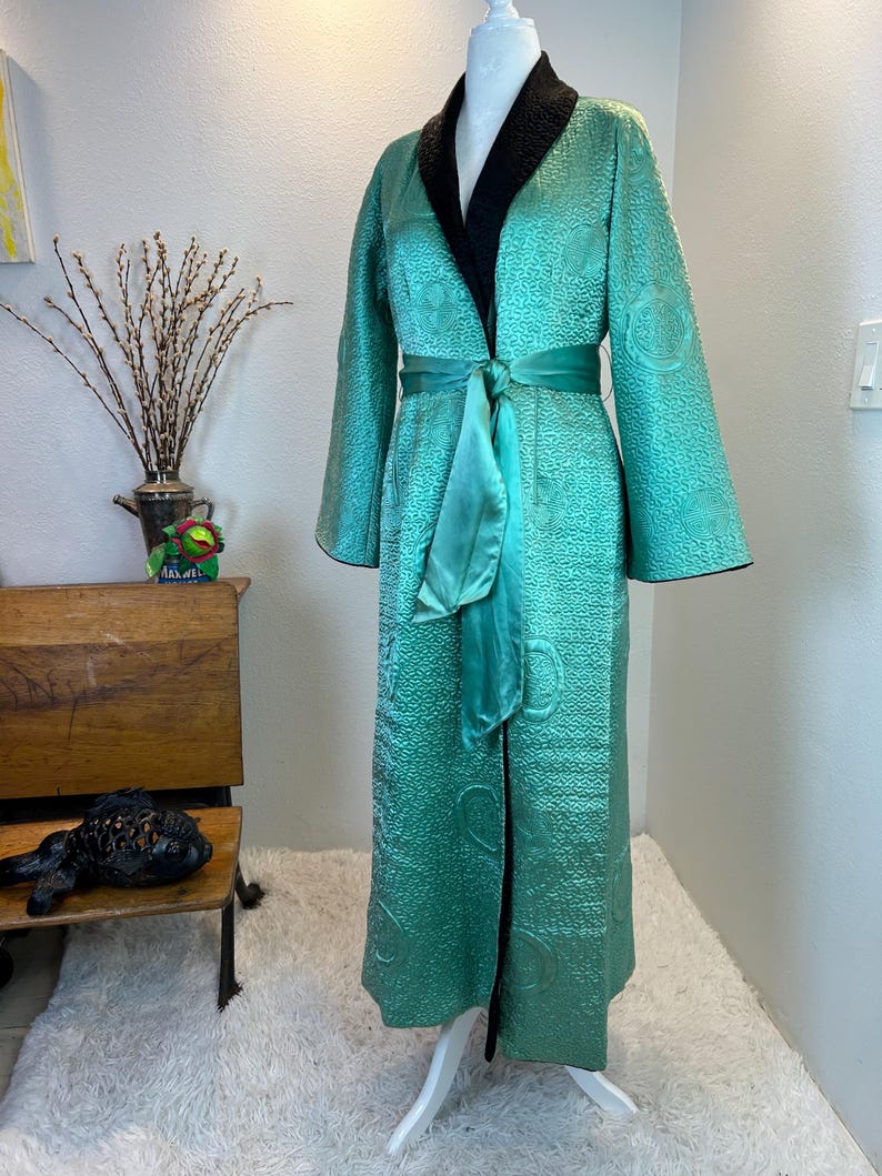 K&ouml;nnte beinhalten: Eine lange, teal-farbene Robe mit strukturiertem Muster und schwarzem Kragen wird auf einer Schaufensterpuppe pr&auml;sentiert. Die Robe hat einen passenden teal-farbenen Sch&auml;rpe, die in der Taille gebunden ist. Die &Auml;rmel sind lang und weit. Es ist ein Vintage-Kleidungsst&uuml;ck.