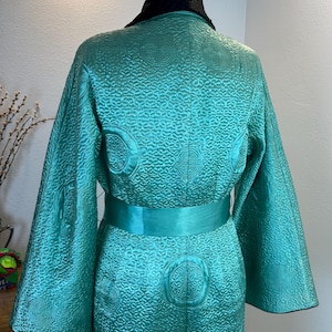 K&ouml;nnte beinhalten: T&uuml;rkisfarbener Robe mit schwarzem Kragen und passendem t&uuml;rkisfarbenem G&uuml;rtel. Die Robe hat ein aufwendiges, gestepptes Muster mit kreisf&ouml;rmigen und geometrischen Designs. Die &Auml;rmel sind weit und flie&szlig;end, und die Robe ist aus einem gl&auml;nzenden Stoff gefertigt.