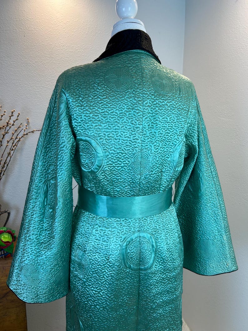 K&ouml;nnte beinhalten: T&uuml;rkisfarbener Robe mit schwarzem Kragen und passendem t&uuml;rkisfarbenem G&uuml;rtel. Die Robe hat ein aufwendiges, gestepptes Muster mit kreisf&ouml;rmigen und geometrischen Designs. Die &Auml;rmel sind weit und flie&szlig;end, und die Robe ist aus einem gl&auml;nzenden Stoff gefertigt.