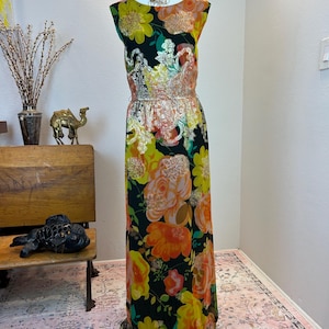 Pode incluir: Um vestido comprido sem mangas com fundo preto e um padrão floral vibrante em tons de laranja, amarelo e verde. O corpete e a cintura são adornados com lantejoulas metálicas douradas e bordados num design de espirais.