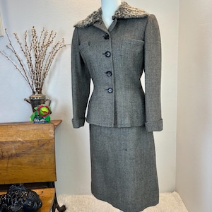 May include: A vintage gray tweed suit displayed on a mannequin. The suit includes a fitted jacket with a faux fur collar and a matching knee-length skirt. The jacket has black buttons and a small pocket. The suit is a classic style.