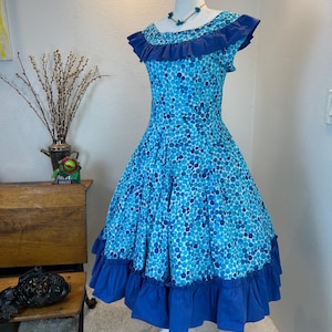 Puede incluir: Vestido vintage de lunares azules sobre un maniquí blanco. El vestido presenta un escote y un dobladillo con volantes, con un corpiño ajustado y una falda acampanada. Un collar floral azul a juego está alrededor del cuello del maniquí.