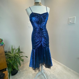 Op de afbeelding: Een blauwe, glinsterende jurk met een strakke buste en een zwierige rok. De rok heeft een kantversiering aan de zoom.