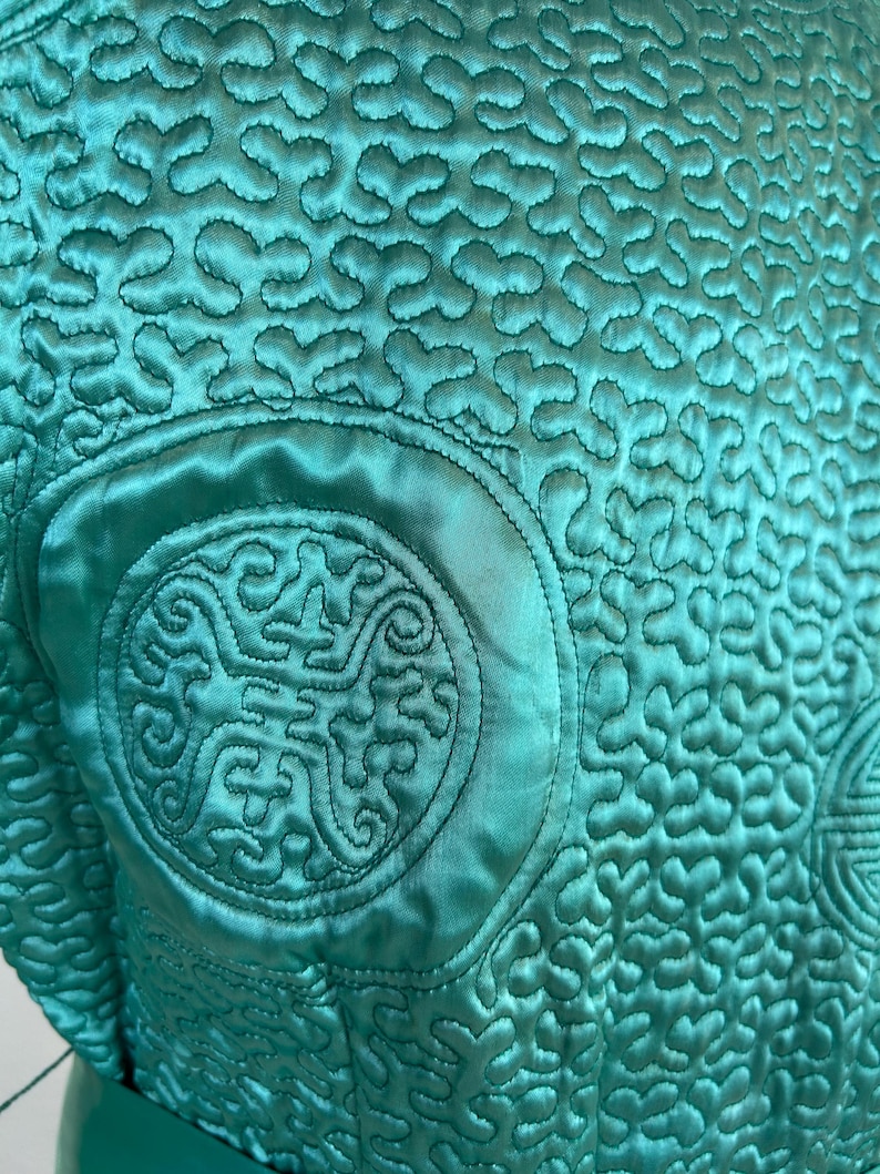 K&ouml;nnte beinhalten: Nahaufnahme eines t&uuml;rkisfarbenen, gesteppten Stoffes mit einem aufwendigen Muster. Der Stoff weist ein sich wiederholendes Design aus kleinen, verbundenen Formen und gr&ouml;&szlig;eren kreisf&ouml;rmigen Stickereidetails auf. Die Textur ist glatt und gl&auml;nzend, was auf ein Satin- oder Seidenmaterial hindeutet.