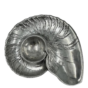May include: A silver-colored, nautilus-shaped serving dish. The dish features a central, rounded bowl and a larger, open area, all with a textured, ridged design. The overall shape resembles a seashell.