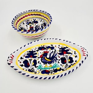 May include: A ceramic bowl and an oval platter featuring hand-painted designs of a blue rooster with a red comb, surrounded by dark blue floral motifs and yellow and red decorative bands on a white background with blue rim accents.