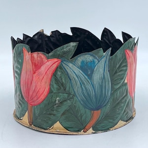 May include: A round, hand-painted container with a floral design. The container features red and blue tulip-like flowers with green leaves. The rim is black and the base is gold. The container is likely made of metal or a similar material.