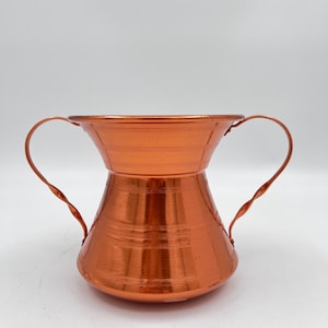 May include: A copper pitcher with two handles. The pitcher has a wide mouth and a narrow base. It has a hammered finish and a shiny, polished look.