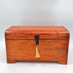 May include: A rectangular wooden chest with a curved top and a dark metal clasp. The chest is made of reddish-brown wood and has a small, decorative tassel hanging from the clasp. The chest sits on four small feet.
