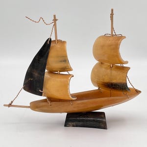 May include: A small, handcrafted sailboat made from horn. The sails are a light brown color, and the hull is a darker brown. The sailboat is mounted on a black horn base.
