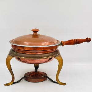 May include: A copper chafing dish with a matching lid and a long, turned wooden handle. The dish rests on a brass-toned metal stand featuring three curved legs and a central circular base designed to hold a heat source beneath the copper bowl.
