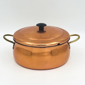 May include: A copper-colored cooking pot with a lid and two handles. The lid has a black knob. The handles are gold-colored. The pot is round and sits on a white surface. The pot is likely used for cooking.