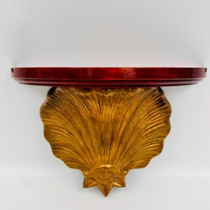May include: A wall-mounted shelf featuring a semi-circular top surface with a deep red, polished finish. The support bracket is shaped like a large, textured gold-colored seashell with radiating ridges and a decorative base element.