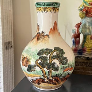 May include: A decorative white ceramic vase with a landscape painting. The vase features a detailed scene of mountains, trees, and a small house. The top of the vase has a green and yellow geometric design.