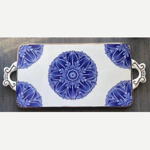 May include: A rectangular white ceramic serving tray featuring a large blue floral mandala pattern in the center and smaller matching blue floral designs in each corner. The tray includes two ornate white handles with gold-colored trim.