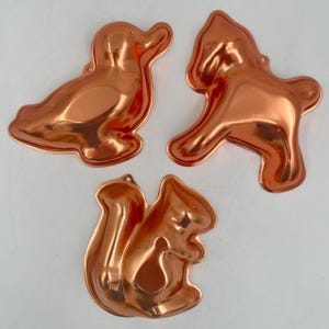 May include: Three copper-colored metal molds in the shapes of a duck, a horse, and a squirrel. Each mold has a loop at the top for hanging. The duck is lying down, the horse is rearing up, and the squirrel is sitting.
