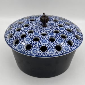 May include: A ceramic flower frog with a dark blue base and a blue and white floral patterned top. The top has numerous holes for arranging flowers and a small, dark brown finial. The design is a classic, elegant style.