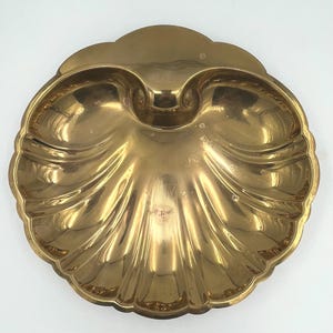 May include: A decorative brass seashell-shaped dish with a scalloped edge. The dish has two compartments and a central indentation. The brass has a polished, reflective surface, and the dish is set against a white background.