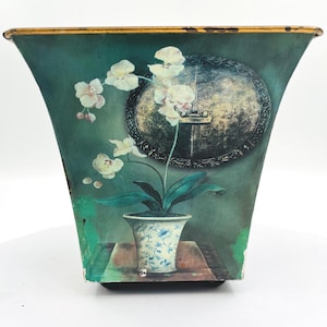 May include: A tapered metal planter with a dark teal finish and a gold-toned rim. The front features a printed illustration of a white orchid in a blue and white patterned pot, set on a table in front of a large, dark, circular decorative wall plaque.