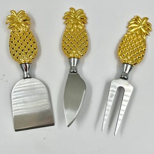 May include: Set of three gold pineapple cheese knives with stainless steel blades. The knives include a spreader, a knife, and a fork.