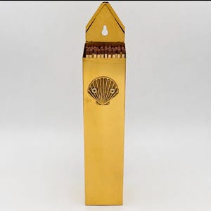 May include: A tall, rectangular, polished brass match holder featuring a pointed top with a keyhole-shaped mounting slot. The front surface is decorated with a centered, embossed scallop shell motif. The open top reveals a row of wooden matches.