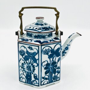 May include: A white and blue ceramic teapot with a brass handle. The teapot features a hexagonal shape and is decorated with blue floral patterns. The lid is also decorated with blue floral designs. The spout is curved and has a floral design.