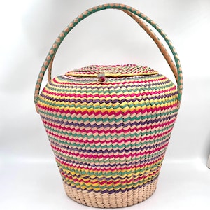 May include: A colorful woven basket with a lid and a braided handle. The basket features horizontal stripes of pink, yellow, green, and purple. The handle is green and tan. A small, red gem is on the lid.
