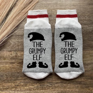 May include: A pair of gray and white socks with red stripes at the top. The socks have a black elf design and the text "The Grumpy Elf" on them.