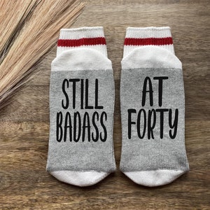 May include: A pair of gray socks with red and white stripes at the top. One sock says "Still Badass" and the other says "At Forty".