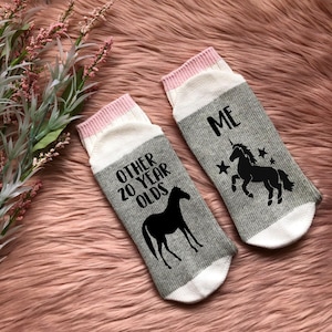 May include: A pair of gray and white socks with pink trim. One sock has a black horse and the text "Other 20 Year Olds". The other sock has a black unicorn and the text "Me".