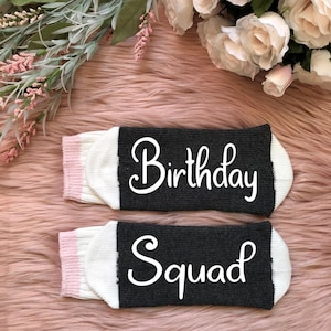 May include: Two pairs of socks with the words "Birthday Squad" in white script. The socks have a dark gray top with white and pink accents. The socks are displayed on a pink, fluffy surface with floral decorations.