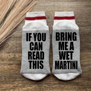 May include: A pair of gray socks with white and red stripes at the top. One sock says "If you can read this" and the other sock says "Bring me a wet martini".