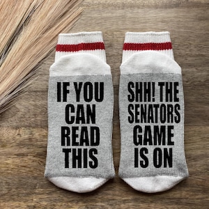 May include: A pair of gray socks with white and red stripes at the top. One sock says "If you can read this" and the other sock says "Shh! The Senators game is on".