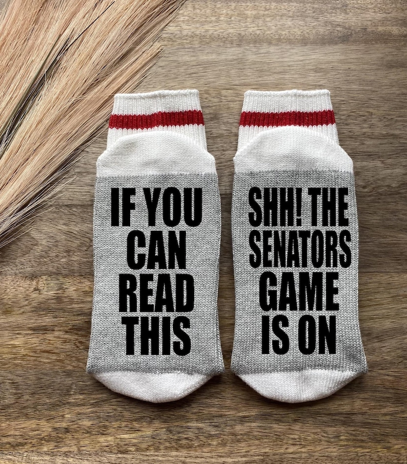 May include: A pair of gray socks with white and red stripes at the top. One sock says "If you can read this" and the other sock says "Shh! The Senators game is on".
