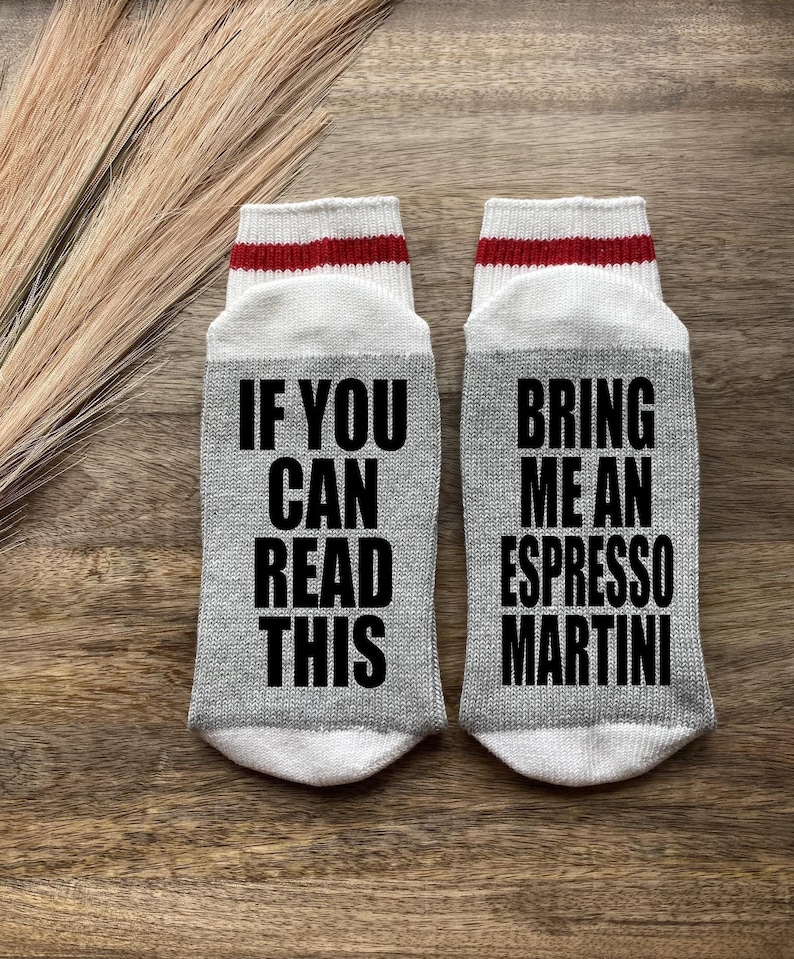May include: A pair of gray socks with white and red stripes. One sock says "If you can read this" and the other sock says "Bring me an espresso martini".