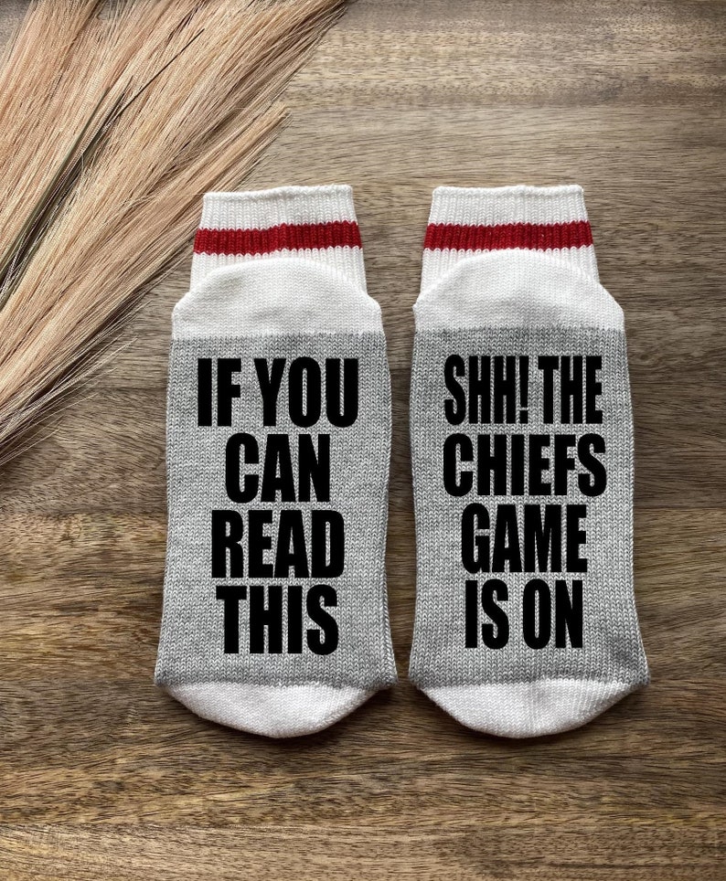 May include: A pair of gray socks with white and red stripes. One sock says "If you can read this" and the other sock says "Shh! The Chiefs game is on".