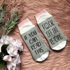 May include: A pair of gray and white socks with pink trim. One sock says "If you can read this" and the other sock says "F*ck off I'm reading".