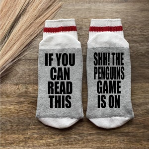 May include: A pair of gray socks with white and red stripes. One sock says "If you can read this" and the other sock says "Shh! The penguins game is on".