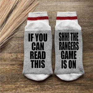 May include: A pair of gray socks with white and red stripes. One sock says "If you can read this" and the other sock says "Shh! The Rangers game is on".