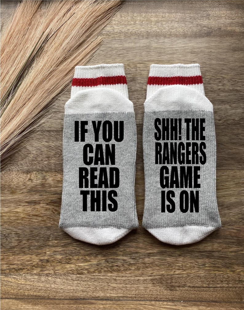 May include: A pair of gray socks with white and red stripes. One sock says "If you can read this" and the other sock says "Shh! The Rangers game is on".