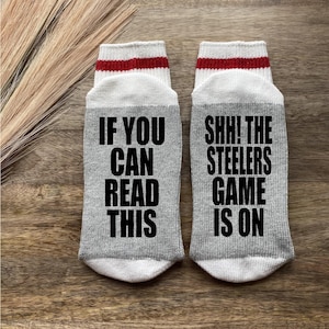 May include: A pair of gray socks with white and red stripes at the top. One sock says "If you can read this" and the other sock says "Shhh! The Steelers game is on".