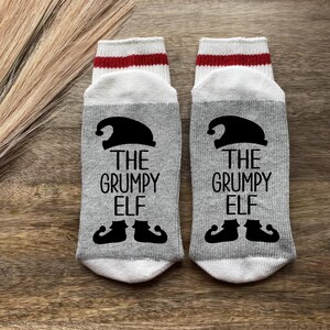 May include: A pair of gray and white socks with red stripes. The socks have a black elf hat and the text "The Grumpy Elf" on them.