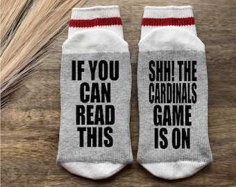 Puede incluir: Un par de calcetines grises con rayas blancas y rojas en la parte superior. Un calcetín dice "Si puedes leer esto" y el otro calcetín dice "¡Shh! El partido de los Cardinals está en marcha".