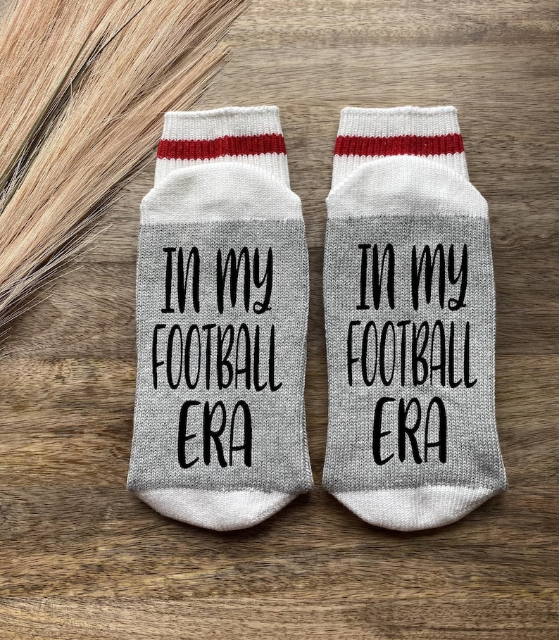 K&ouml;nnte beinhalten: Ein Paar graue Socken mit roten und wei&szlig;en Streifen oben. Die Socken haben den Text "In my football era" aufgedruckt.