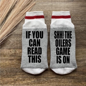 May include: A pair of gray socks with white and red stripes. One sock says "If you can read this" and the other sock says "Shh! The Oilers game is on".