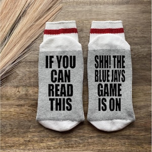 May include: A pair of grey socks with white and red stripes. One sock says "If you can read this" and the other sock says "Shh! The Blue Jays game is on".