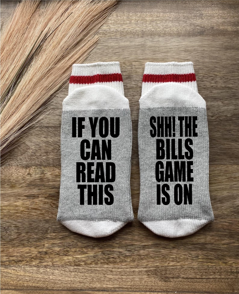 May include: A pair of gray socks with white and red stripes. One sock says "If you can read this" and the other sock says "Shh! The Bills game is on".