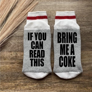 Peut inclure: Une paire de chaussettes grises avec des rayures rouges et blanches en haut. Une chaussette dit "Si vous pouvez lire ceci" et l'autre chaussette dit "Apportez-moi un coca".