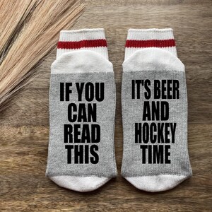 May include: A pair of gray socks with white and red stripes at the top. One sock says "If you can read this" and the other sock says "It's beer and hockey time".