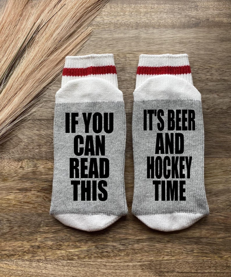 May include: A pair of gray socks with white and red stripes at the top. One sock says "If you can read this" and the other sock says "It's beer and hockey time".
